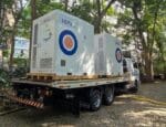 Sistema BESS da Ion Energia sendo transportado em caminhão, instalado em área arborizada com edifícios ao fundo.