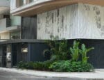 Fachada de edifício contemporâneo com revestimento metálico perfurado branco, destacando padrões geométricos e vegetação paisagística na entrada.