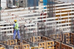 Trabalhador da construção civil com equipamentos de segurança inspeciona estrutura de concreto e aço em obra.