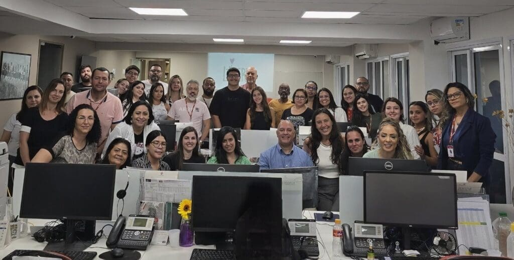 Colaboradores da MBigucci reunidos em um escritório durante o lançamento do Programa Big Escudo, todos sorrindo e demonstrando engajamento com a iniciativa de proteção à infância.