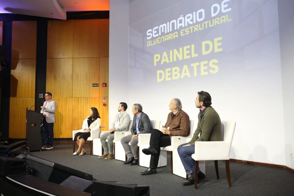 Painel de debates do Seminário de Alvenaria Estrutural com seis especialistas no palco, incluindo uma mulher, enquanto um moderador conduz a discussão ao púlpito, diante de um telão com o nome do evento.