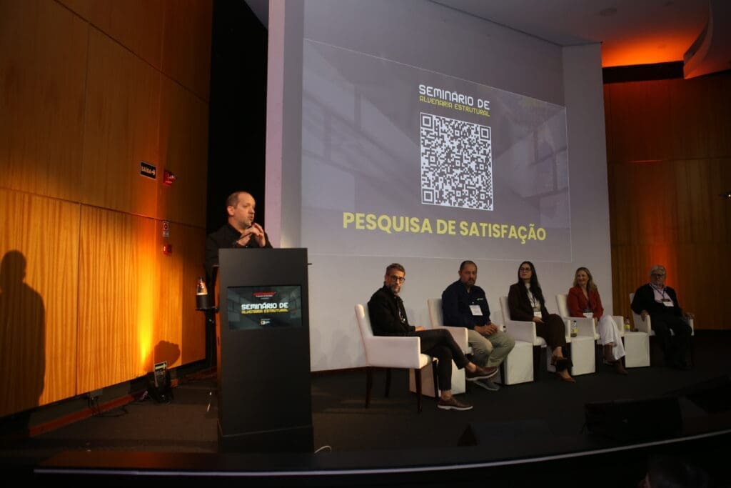 Painel de especialistas no palco do Seminário de Alvenaria Estrutural, com moderador ao púlpito e projeção de QR Code para pesquisa de satisfação ao fundo.