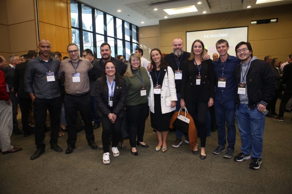 Grupo de profissionais da construção civil posa para foto durante evento de networking, com crachás de identificação e ambiente corporativo ao fundo.
