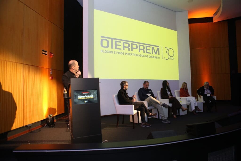 Logo da Oterprem projetado em telão durante apresentação de vídeo institucional no Seminário de Alvenaria Estrutural, com painel de debatedores no palco.