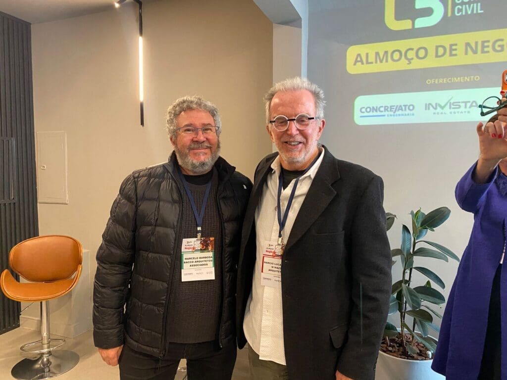 Dois profissionais do setor da construção civil sorrindo juntos durante o 65° Almoço de Negócios do C3, com crachás de identificação e painel do evento ao fundo.