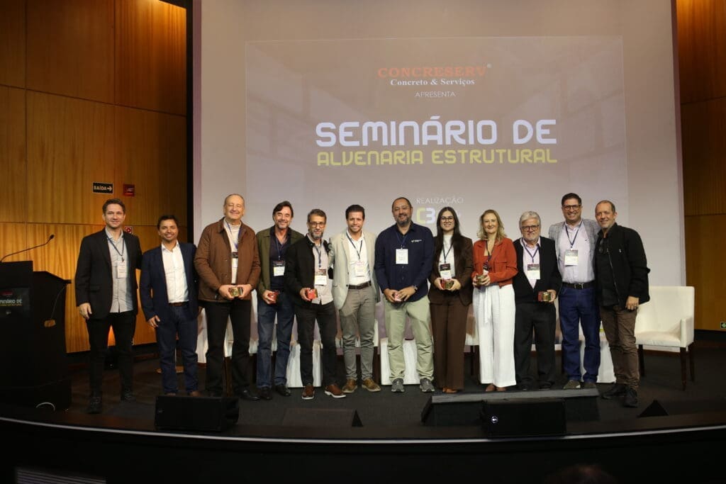 Palestrantes e organizadores do Seminário de Alvenaria Estrutural do C3 posam juntos no palco após o evento, segurando troféus de reconhecimento, diante do telão com o nome do seminário.
