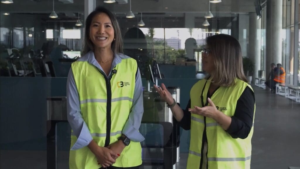 Bárbara Martinez, gerente de Relacionamentos do C3 - O Clube da Construção Civil, e Kristina Wu, gerente comercial técnica da Guardian Glass, conversam sorrindo durante visita técnica ao Beyond The Club São Paulo, ambas usando coletes de segurança amarelos.