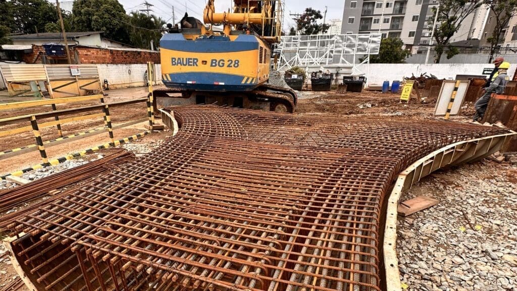 Armação de aço curvada para fundação em obra de construção civil, com escavadeira ao fundo e trabalhadores no canteiro.