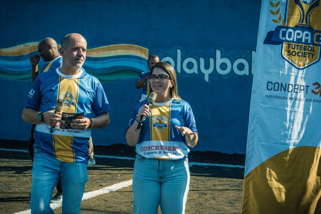 Rodolfo Zagallo, CEO do C3, e Bárbara Martinez, Gerente de Relacionamento do C3, apresentam a abertura oficial do Torneio Integração da Copa C3 2026, usando uniformes do evento e microfones em mãos, em frente ao banner da competição.