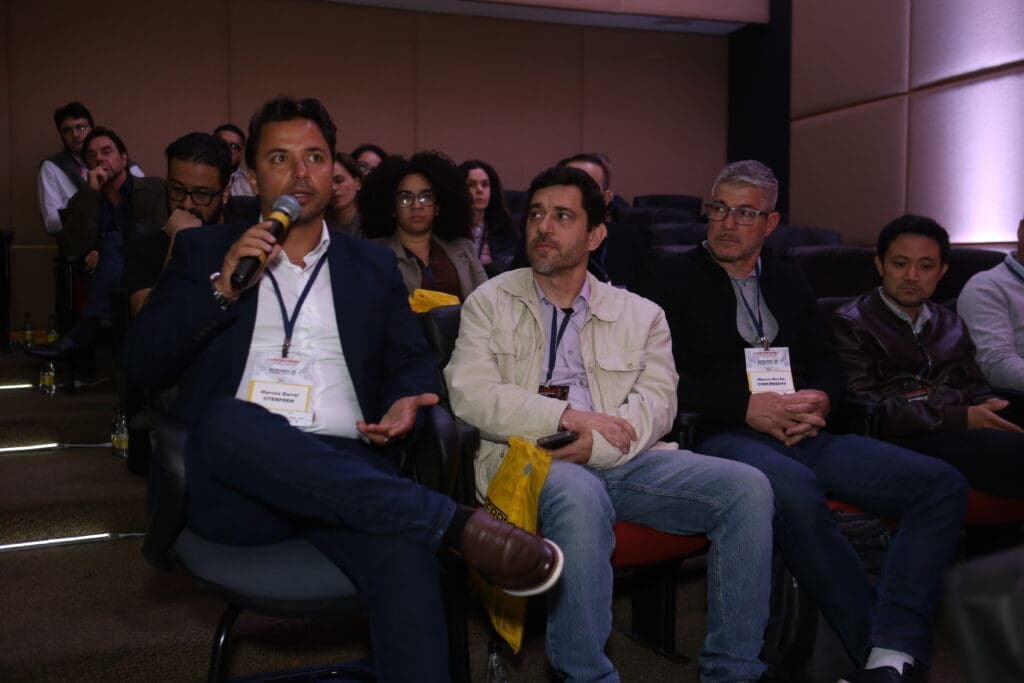 Marcos Barral, Diretor Comercial da Oterprem, faz uma intervenção com microfone durante painel de debates, sentado entre outros participantes em auditório, com plateia atenta ao fundo.