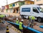 Equipe de trabalhadores da Concrejato Engenharia instala tubulação de saneamento básico em rua urbana, utilizando equipamentos modernos e seguindo rigorosos protocolos de segurança, com vans da empresa ao fundo e casas populares ao redor.