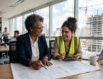 Duas engenheiras, uma mais experiente e outra jovem, analisam plantas de obra e sorriem em um escritório moderno com vista para um canteiro de construção civil ao fundo.