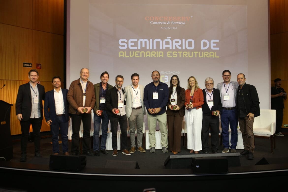 Grupo de palestrantes e organizadores do Seminário de Alvenaria Estrutural do C3 posam juntos no palco do auditório, com o telão ao fundo exibindo o nome do evento.