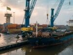 Navio cargueiro carregado de aço sendo descarregado por guindastes em porto brasileiro, com bandeira do Brasil ao fundo ao amanhecer