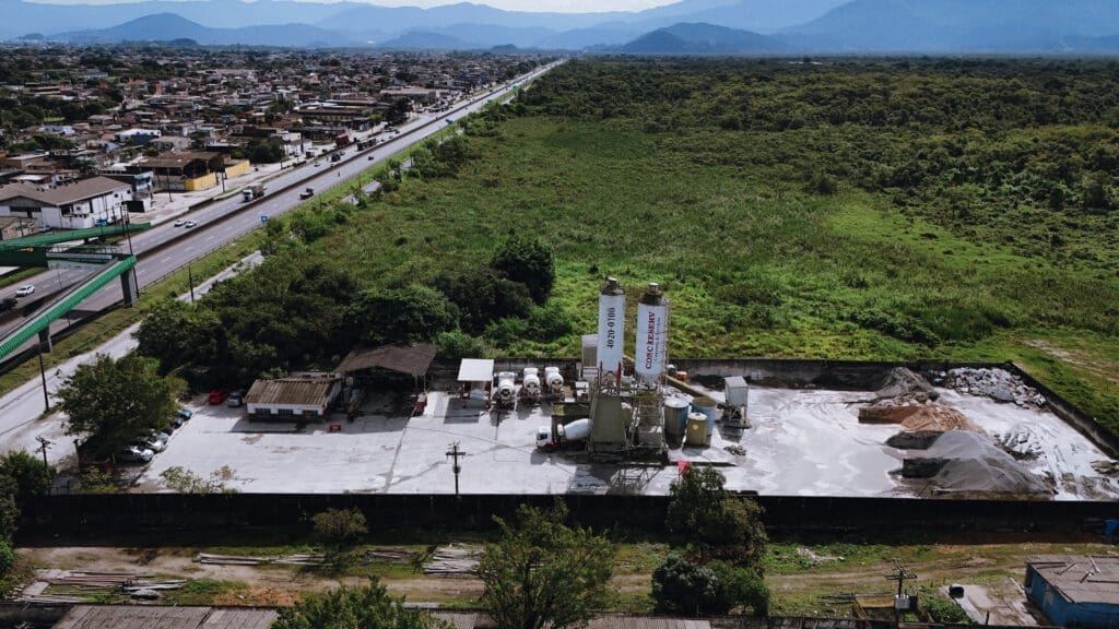 Vista aérea da filial Guarujá da Concreserv, mostrando a unidade de concreto usinado ao lado de uma rodovia movimentada e áreas verdes, com a cidade e montanhas ao fundo.