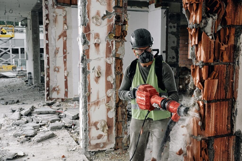 Operador utiliza martelete demolidor Hilti TE 600-AVR em demolição de parede de tijolos, equipado com EPI completo em ambiente de obra.