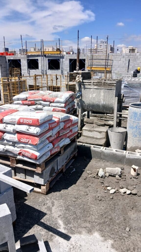 Sacos de argamassa estrutural AAE8 empilhados em paletes em um canteiro de obras, com paredes de blocos em construção ao fundo.