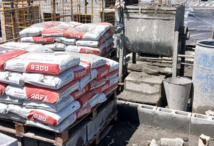 Sacos de argamassa estrutural AAE8 empilhados em paletes em um canteiro de obras, com paredes de blocos em construção ao fundo.