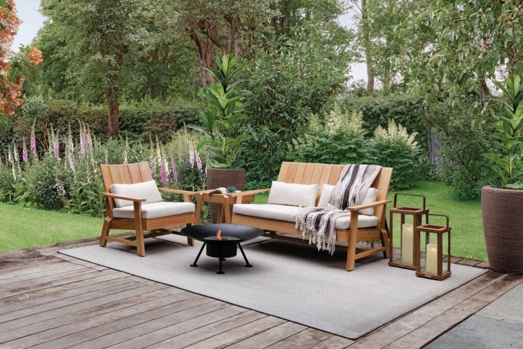 Sofá de dois lugares, poltrona e mesa lateral da linha Marco da Tidelli, em madeira com almofadas claras, dispostos em um jardim ao ar livre sobre tapete, com lanternas decorativas e vegetação ao fundo.