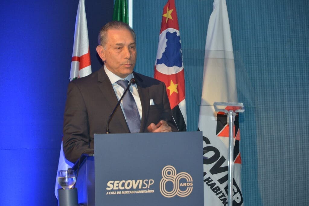 Jorge Cury Neto, novo presidente do Secovi-SP para o biênio 2026-2028, discursa na cerimônia de posse, ao lado de bandeiras e púlpito institucional.