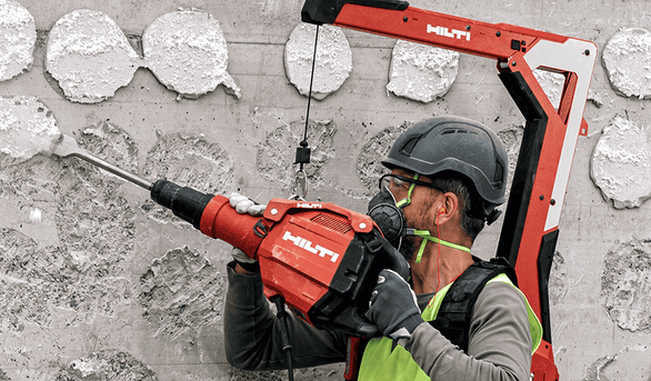 Trabalhador utiliza martelete demolidor Hilti TE 600-AVR com exoesqueleto EXO-T-22 para remover revestimento de parede de concreto, equipado com EPI completo.