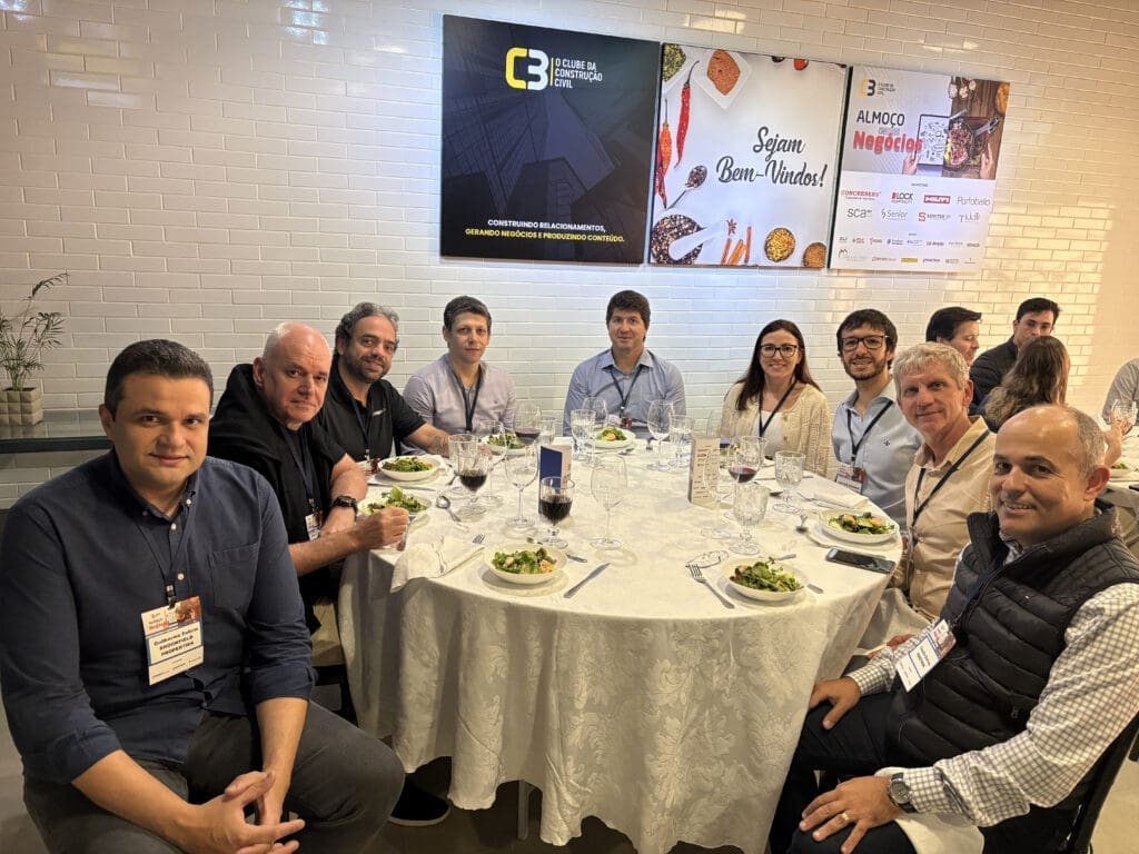 Grupo de profissionais da construção civil reunidos ao redor de uma mesa redonda durante o 66° Almoço de Negócios do C3, com crachás de identificação, pratos servidos e painéis institucionais ao fundo.