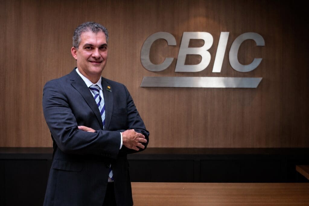 Homem de terno escuro, sorrindo, posando de braços cruzados em frente ao logo da CBIC em uma parede de madeira.