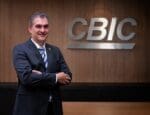 Homem de terno escuro, sorrindo, posando de braços cruzados em frente ao logo da CBIC em uma parede de madeira.