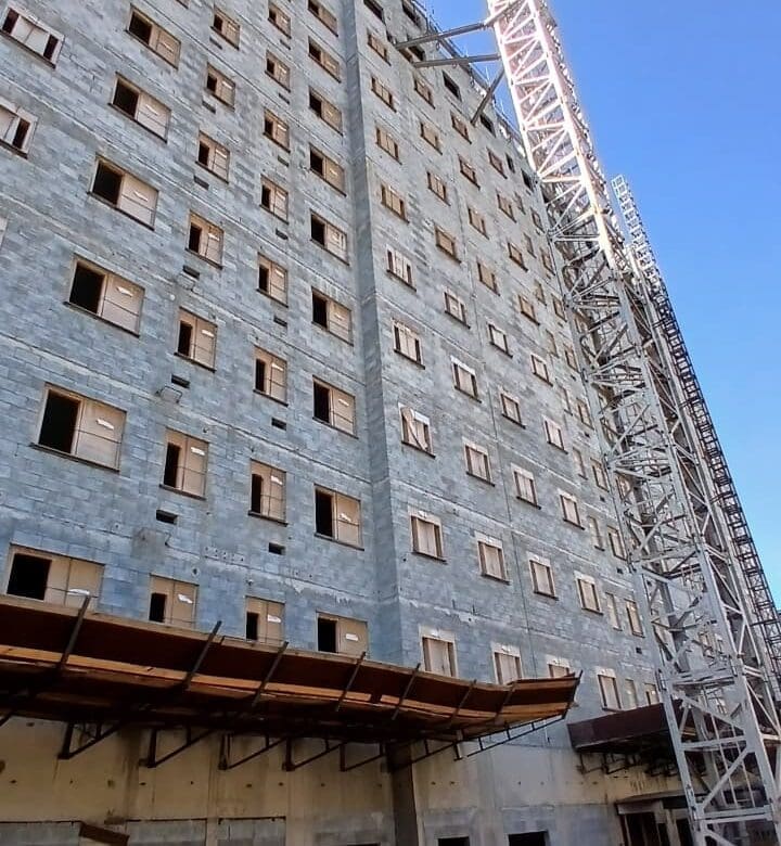 Edifício alto em construção com estrutura de alvenaria aparente e guindaste de torre ao lado, sob céu azul.