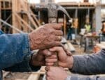 Mãos de trabalhadores de diferentes idades segurando juntas um martelo em um canteiro de obras, simbolizando a passagem de conhecimento entre gerações na construção civil. Imagem Ilustrativa criada pela Equipe C3.