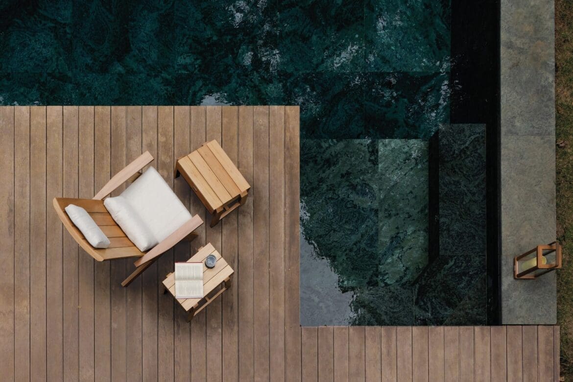Poltrona deck da linha Marco da Tidelli, inspirada na cadeira Adirondack, com puff e mesa lateral em madeira, posicionados sobre deck ao lado de piscina.