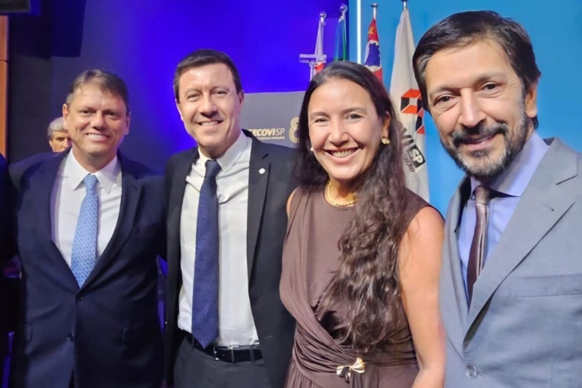 Quatro líderes do setor público e privado posam sorrindo durante a cerimônia de posse da diretoria do Secovi-SP, com bandeiras ao fundo e iluminação azul.