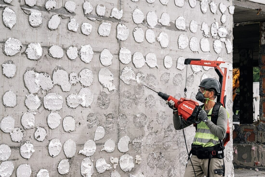 Operador utiliza martelete demolidor Hilti TE 600-AVR com exoesqueleto EXO-T-22 em obra, removendo revestimento de parede de concreto com segurança e ergonomia.